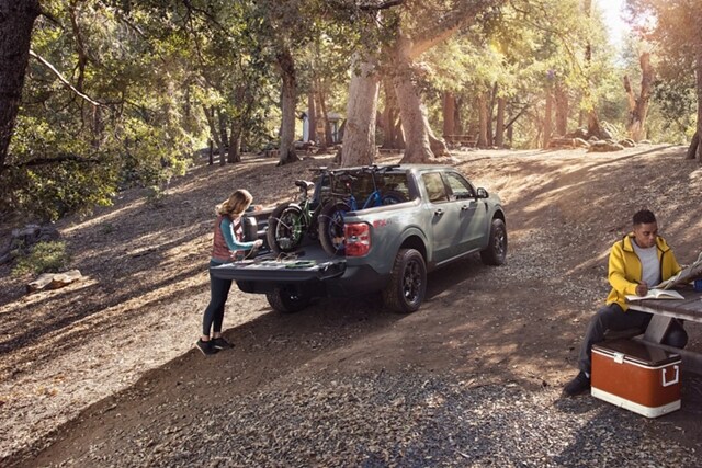Woman at a campsite plugs a bike into the 400-watt 110V outlet in the bed of a 2023 Ford Maverick® XLT model in color Area 51