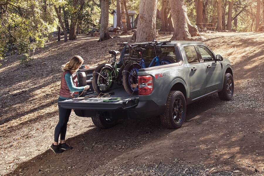 Woman at a campsite plugs a bike into the 400-watt 110V outlet in the bed of a 2023 Ford Maverick® XLT model in color Area 51