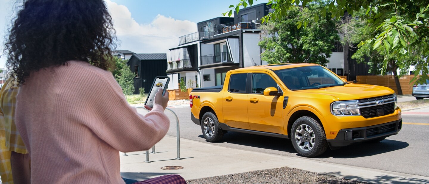 Woman using the FordPass® App on her phone to control a Cyber Orange 2023 Ford Maverick® truck parked on the street