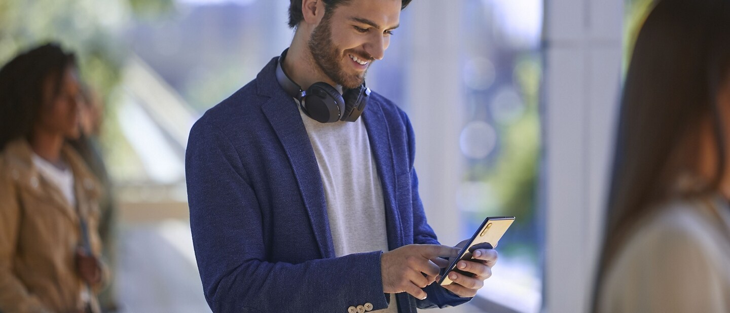 Man with headphones around neck using his phone in a sunny building with people in the background