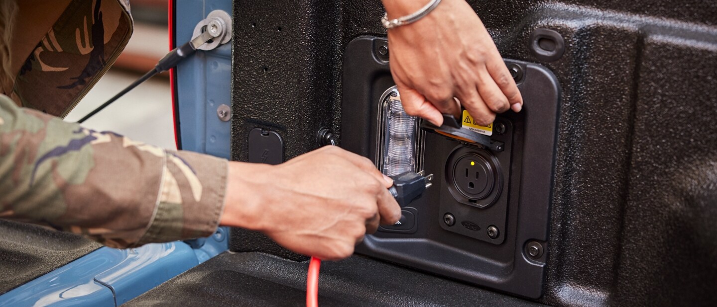 Una mano enchufa un cable de extensión en un tomacorriente de 110 V y 400 vatios en la plataforma de una camioneta Ford Maverick® 2023