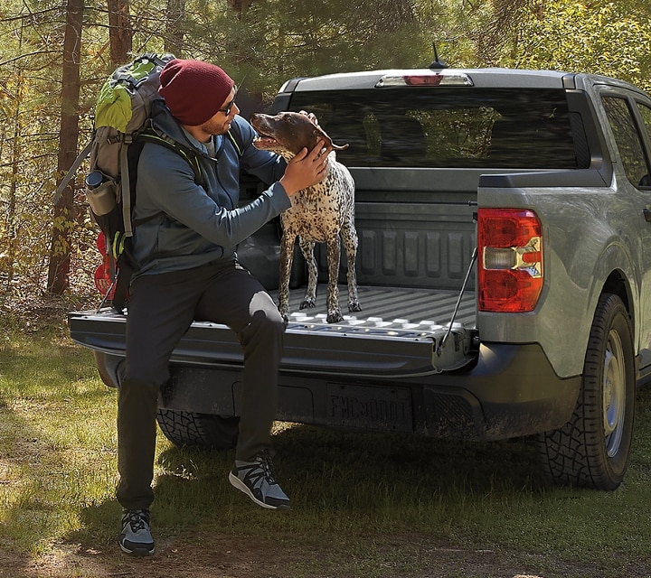 Una camioneta Ford Maverick® 2023 estacionada cerca de un camino de montaña y un hombre y su perro sentados en la puerta trasera