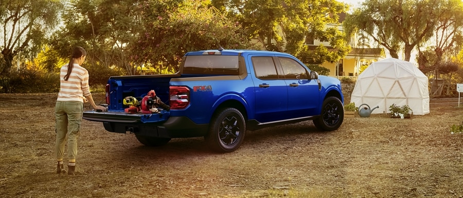 A 2024 Ford Maverick® truck in Atlas Blue Metallic is parked at a campsite with the bed being unloaded by a person