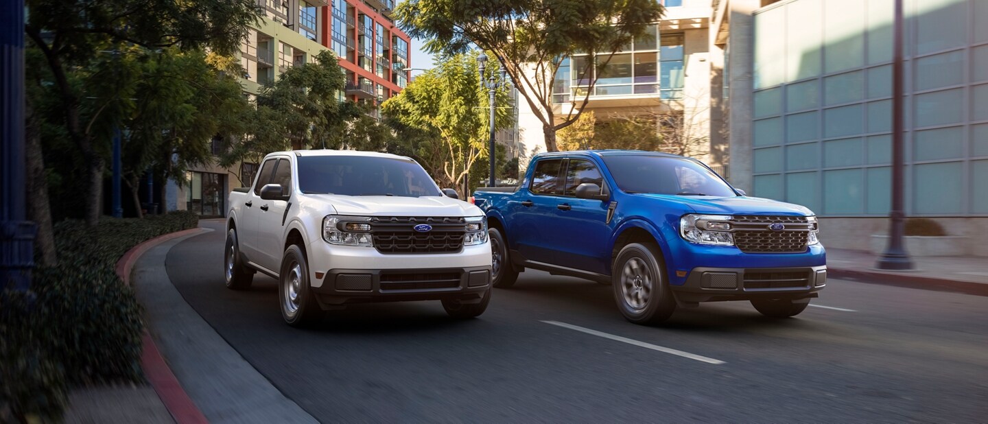 Two 2024 Ford Maverick® trucks in Oxford White and Atlas Blue Metallic are being driven on a tree-lined city road