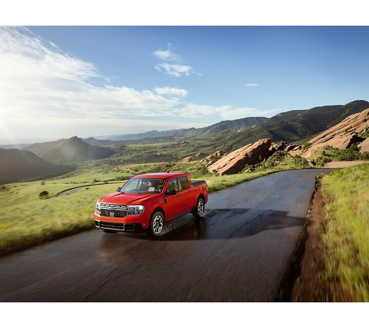 A 2024 Ford Maverick® truck in Hot Pepper Red is being driven on a road in a scenic location with mountains in the background