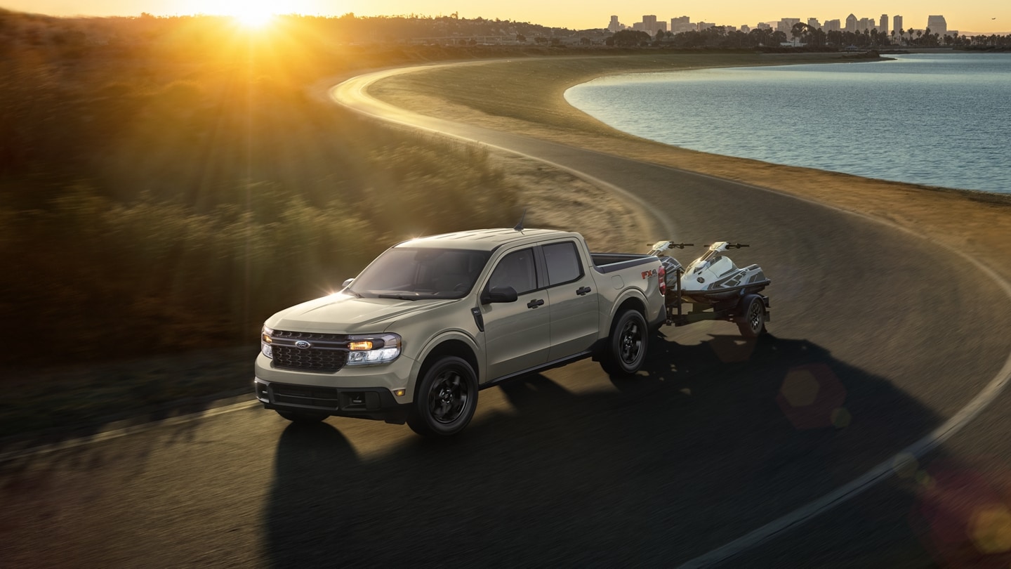 A 2024 Ford Maverick® with jet skis being towed on a city highway at sunset near water