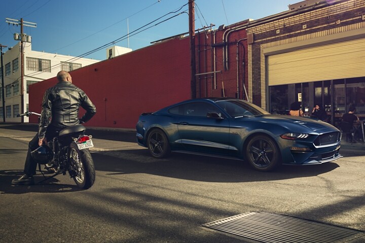 A man on a motorcycle looking at a 2021 Ford Mustang parked on a city street
