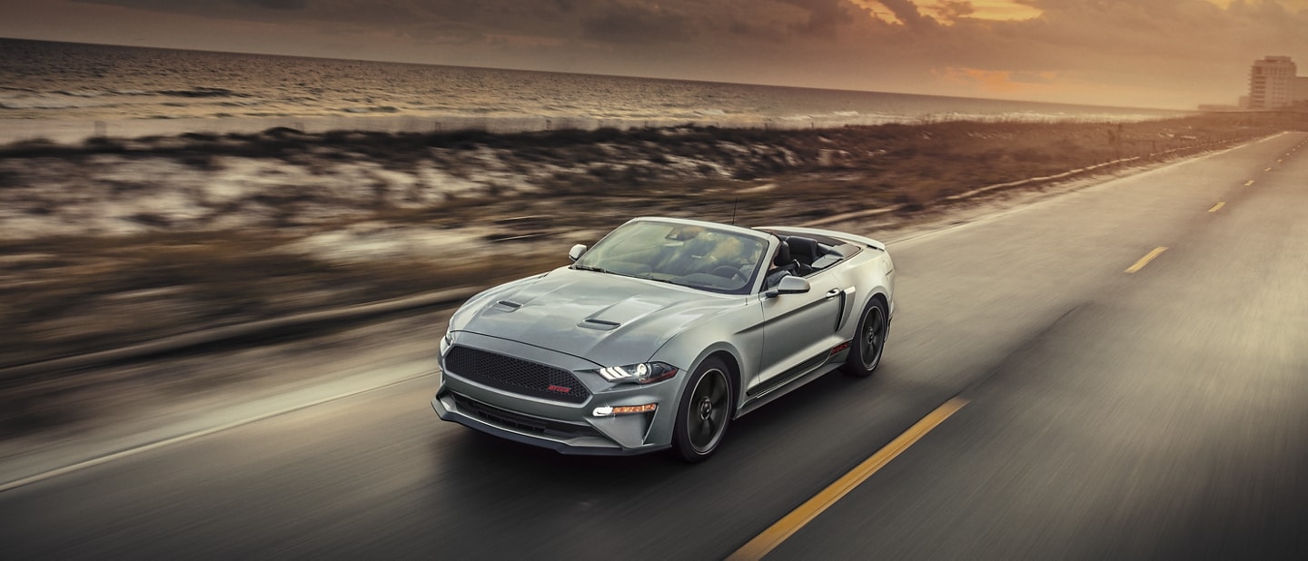 Un Ford Mustang® 2023 convertible con el Paquete California Special, con el techo bajo, circulando por una carretera junto al mar