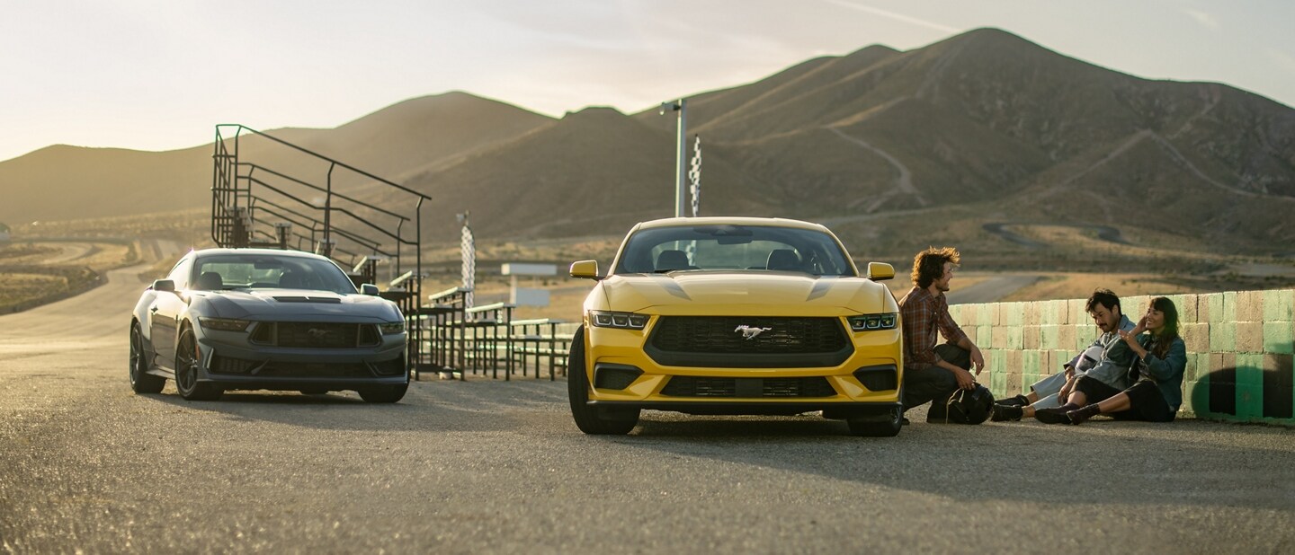 Two 2024 Ford Mustang® coupes parked near a racecourse with three people nearby