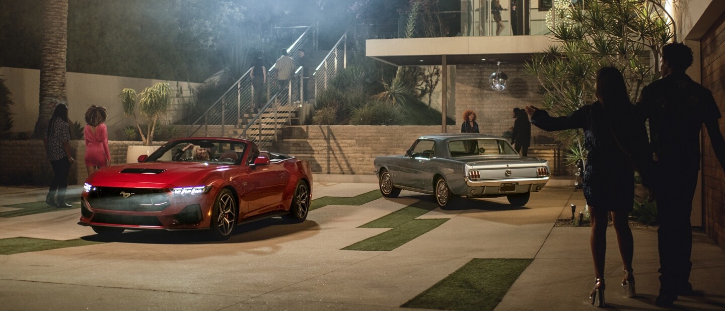 2024 Mustang® GT convertible and a first-gen Mustang coupe parked in a driveway with people nearby
