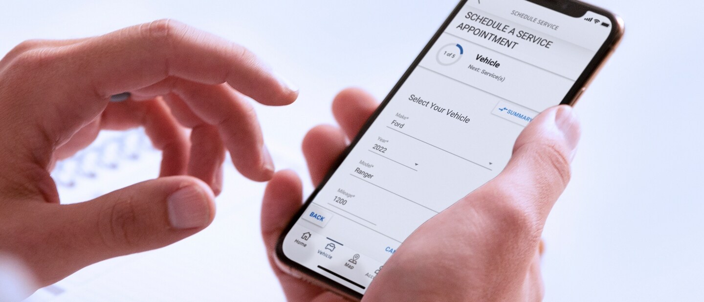 A hand holds a smartphone displaying the Schedule Service menu in the FordPass App
