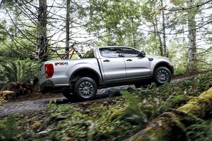 Una Ford Ranger XLT 2023 subiendo una colina en un camino boscoso con accesorio de rieles opcional montado en la plataforma