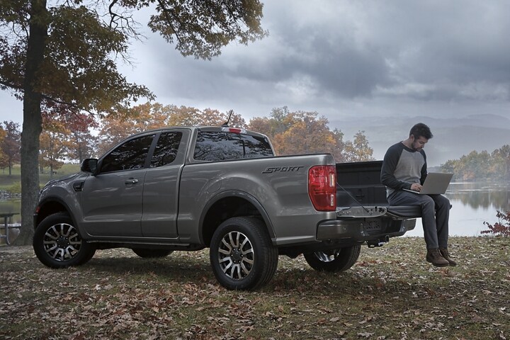 Man sitting on the bed of a 2023 Ford Ranger Lariat Sport, parked near a tree in carbonized grey