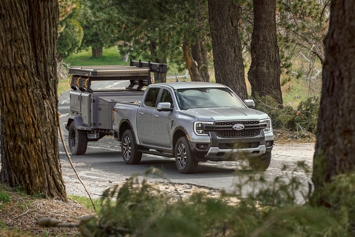 A 2024 Ford Ranger® LARIAT® truck in Iconic Silver is pulling a trailer on a tree-lined road