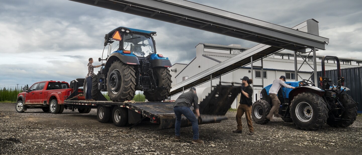 Un hombre cargando un tractor sobre un tráiler enganchado a una Ford Super Duty® F-450® XL SuperCrew®​​​​​​​ Cab 2023 cerca de una fábrica de granos