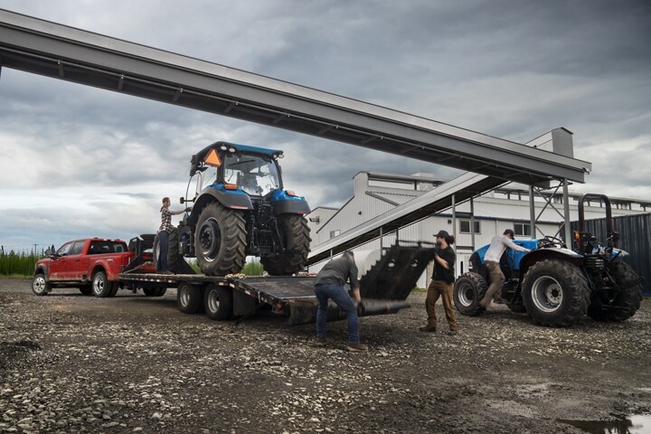 Un hombre cargando un tractor sobre un tráiler enganchado a una Ford Super Duty® F-450® XL Super Crew Cab 2023 cerca de una fábrica de granos
