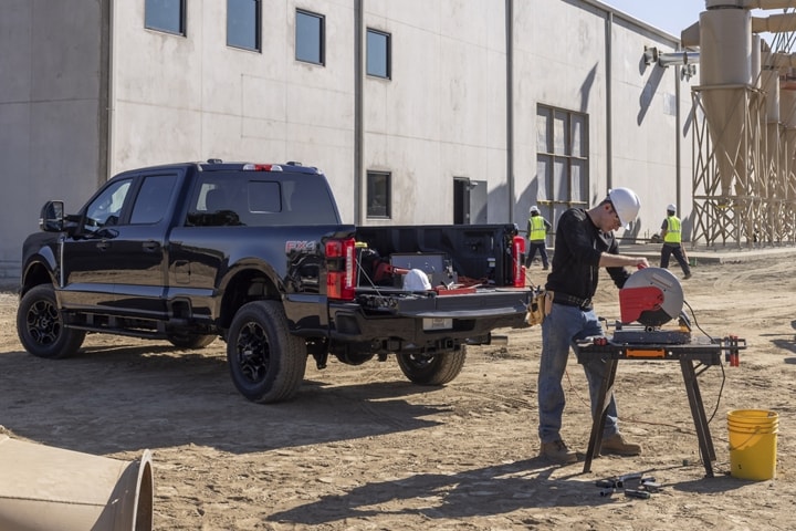 Hombre usando una sierra ingletadora en un lugar de trabajo con una camioneta Ford Super Duty® 2023 estacionada cerca con la puerta trasera baja