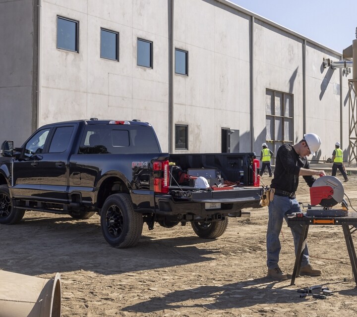 Hombre usando una sierra ingletadora en un lugar de trabajo con una camioneta Ford Super Duty® 2023 estacionada cerca con la puerta trasera baja
