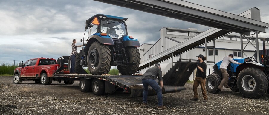 Un hombre cargando un tractor sobre un tráiler enganchado a una Ford Super Duty® F-450® XL SuperCrew®​​​​​​​ Cab 2023 cerca de una fábrica de granos