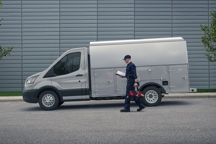 A 2023 Ford Transit® Chassis Cab mechanics truck upfit in front of large building with worker walking towards van