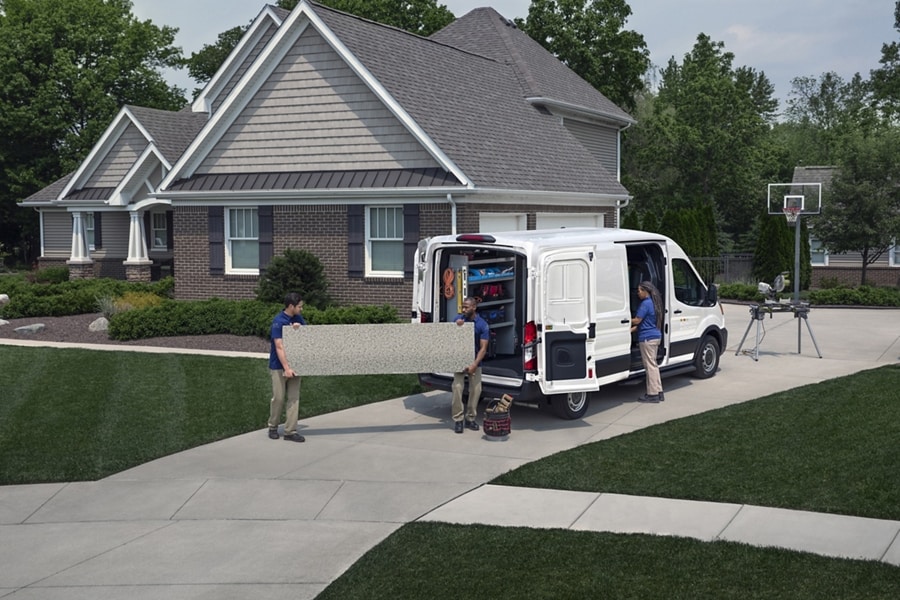 A 2023 Ford Transit® van parked at a home with two men taking a piece of drywall out of the back