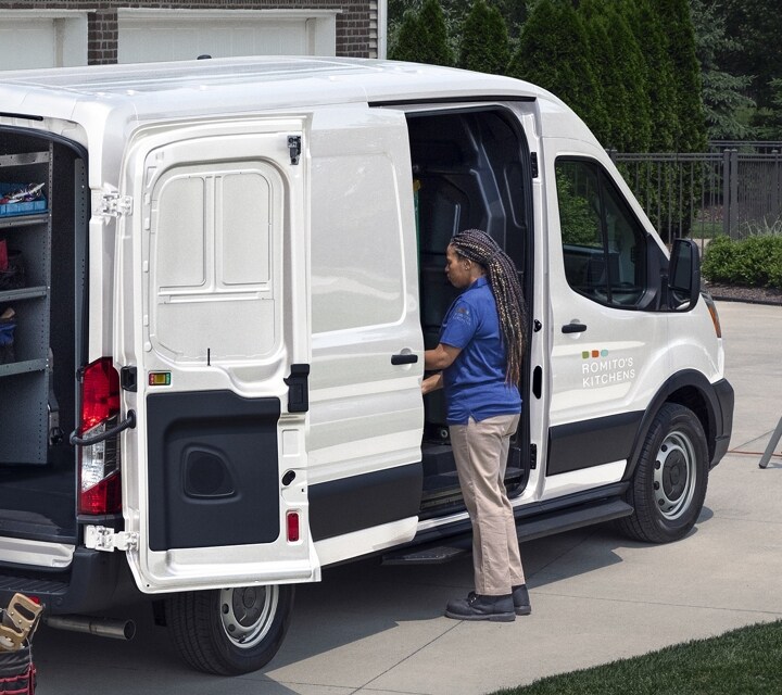 Vista trasera de una Transit® Cargo van 2023 estacionada en una entrada con las puertas de carga lateral y trasera abiertas