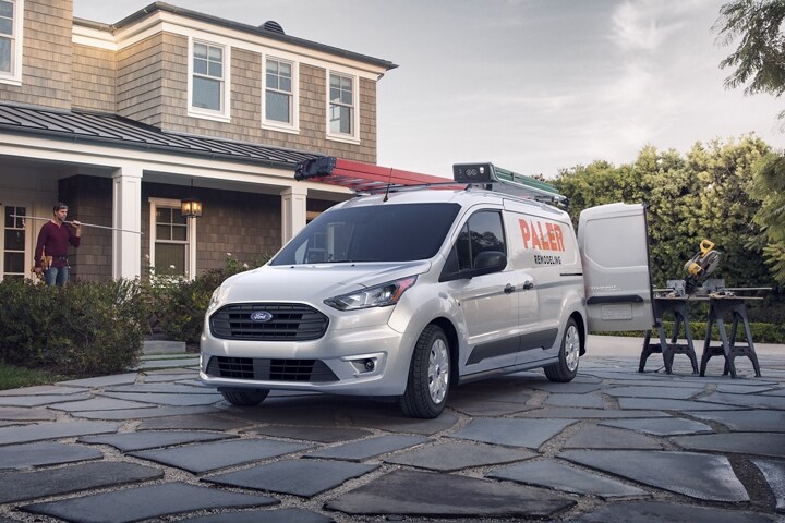 Se muestra una Ford Transit Connect Cargo Van 2023 en Silver con gráficos comerciales no originales disponibles enfrente de una casa