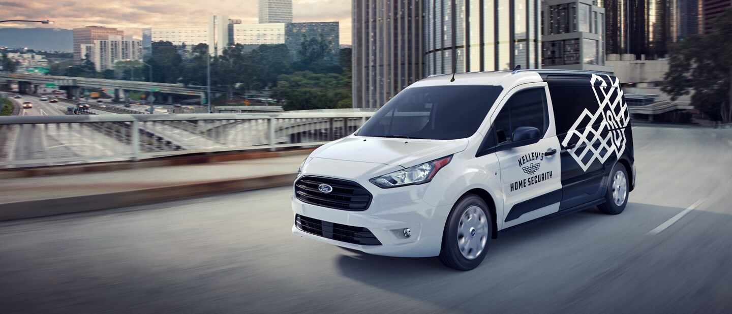 Ford Transit Connect Cargo Van 2023 en Frozen White con gráficos comerciales no originales andando por una calle de ciudad