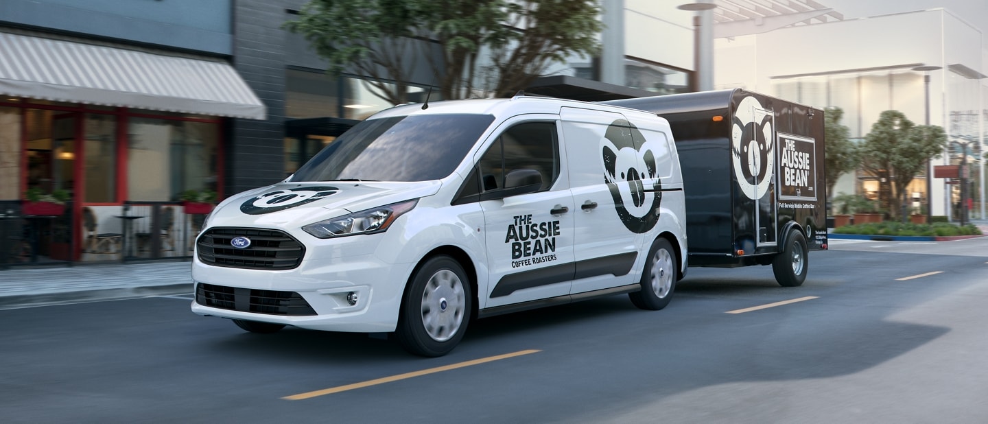 2023 Ford Frozen White Transit Connect Cargo Van with business graphics towing a trailer in the city