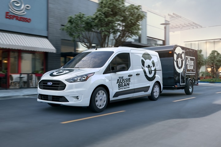2023 Frozen White Transit Connect Cargo Van with available aftermarket business graphics towing a trailer in the city