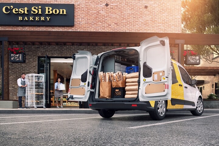 Se muestra una Ford Transit Connect Cargo Van 2023 con puertas de carga traseras de apertura hacia afuera estacionada frente a una panadería