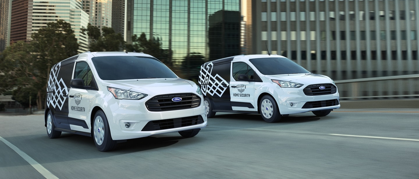 Two 2023 Ford Transit Connect Cargo Vans with eye-catching business graphics being driven on a city street