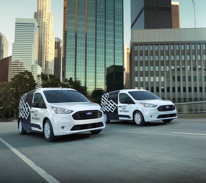 Two 2023 Ford Transit Connect Cargo Vans with aftermarket business graphics being driven on the road