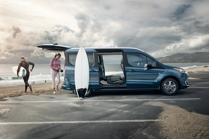 Un Wagon para Pasajeros Ford Transit Connect 2023 en Blue estacionado en la playa con surfistas 