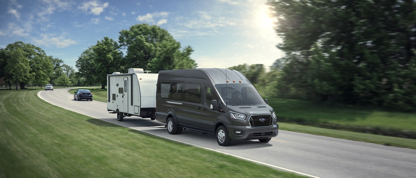 A 2024 Ford Transit® van towing a trailer down a wooded road